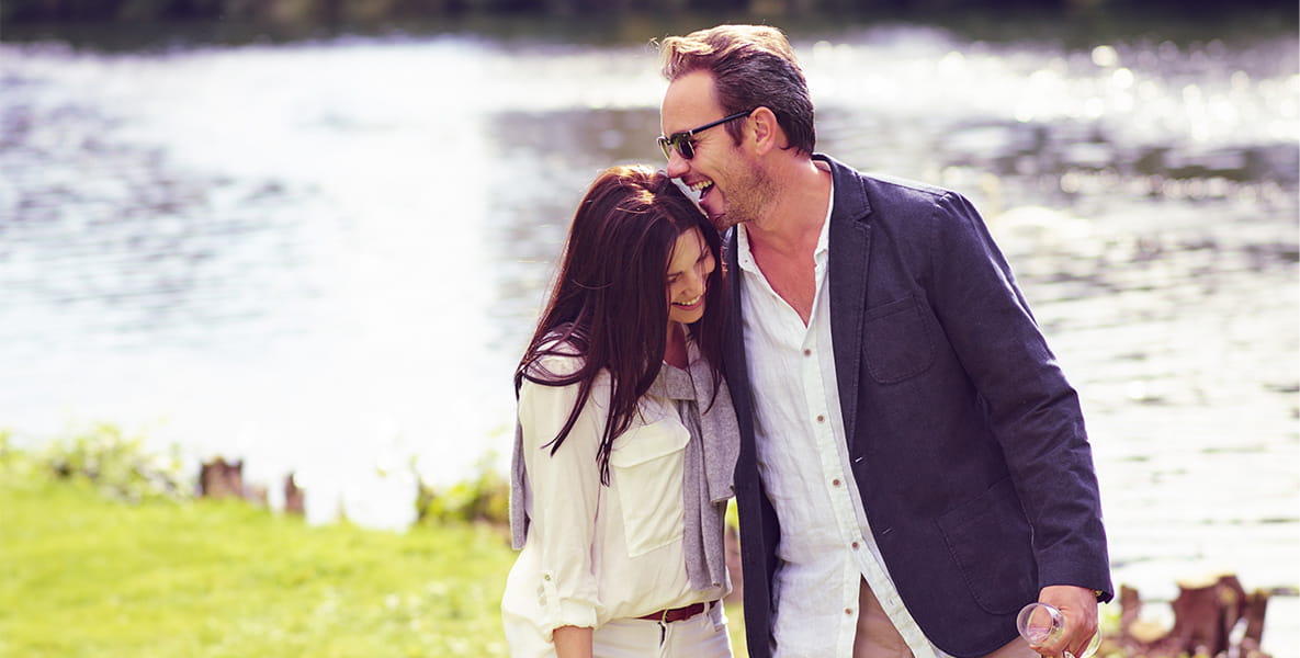 Smiling couple embracing by a lakeside, with the man holding a bottle and glasses on a sunny day.