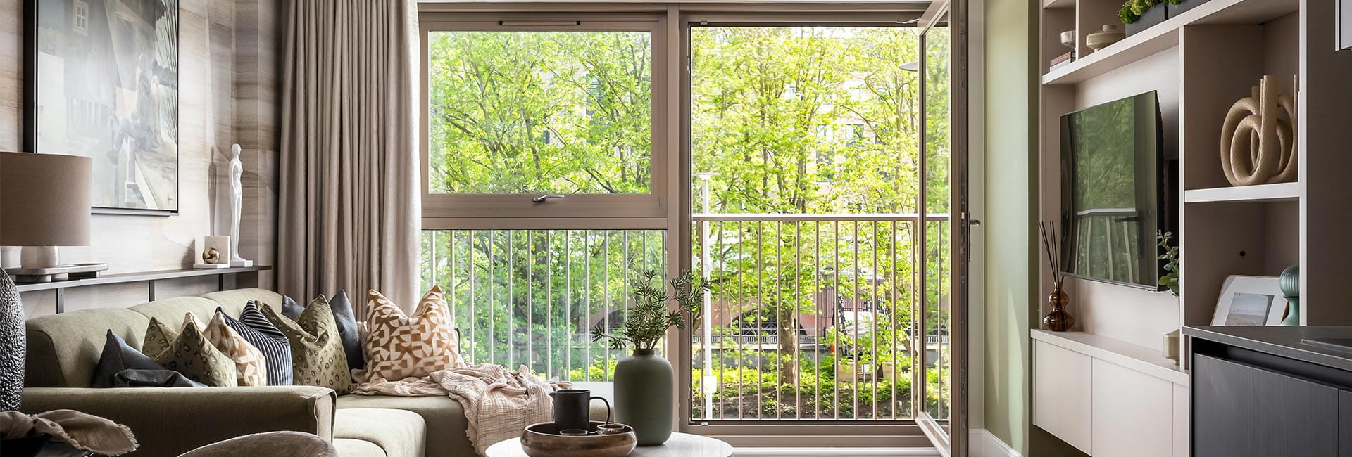 Living space with seating, coffee table and TV unit decorated in beige tones, balcony doors in background with a view of greenery.