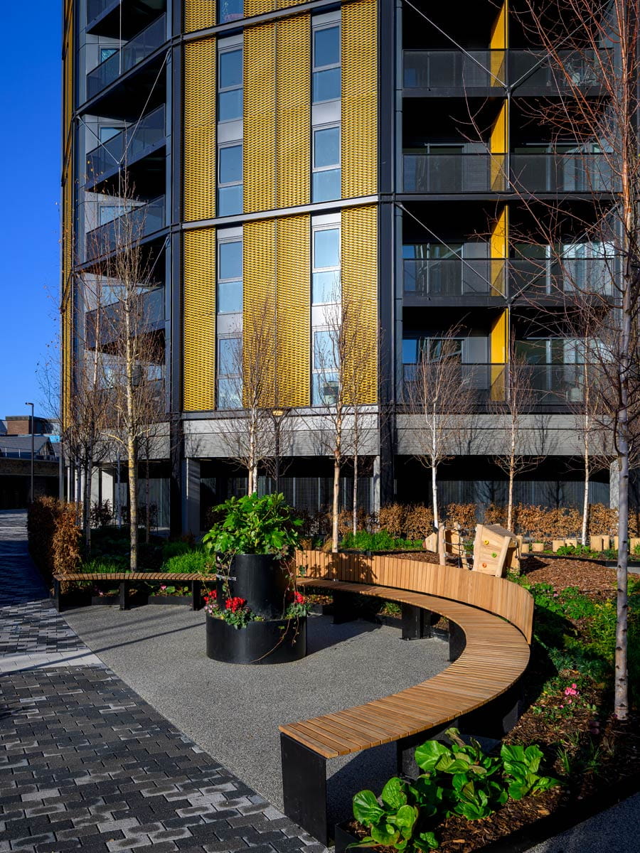Modern apartment building with yellow cladding, balconies, bare trees and curved benches in a landscaped courtyard.