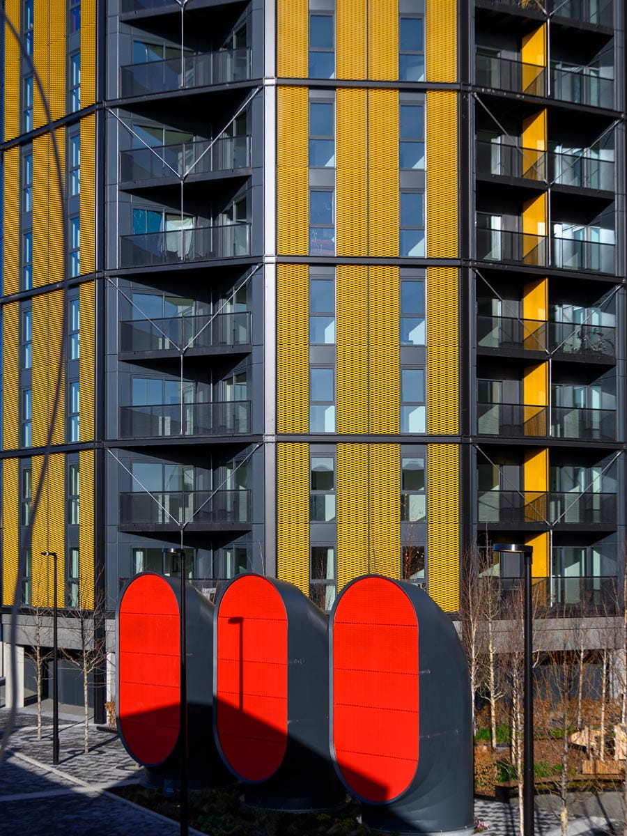 Modern apartment block with yellow cladding, dark balconies and a bold red sculptural structure below.