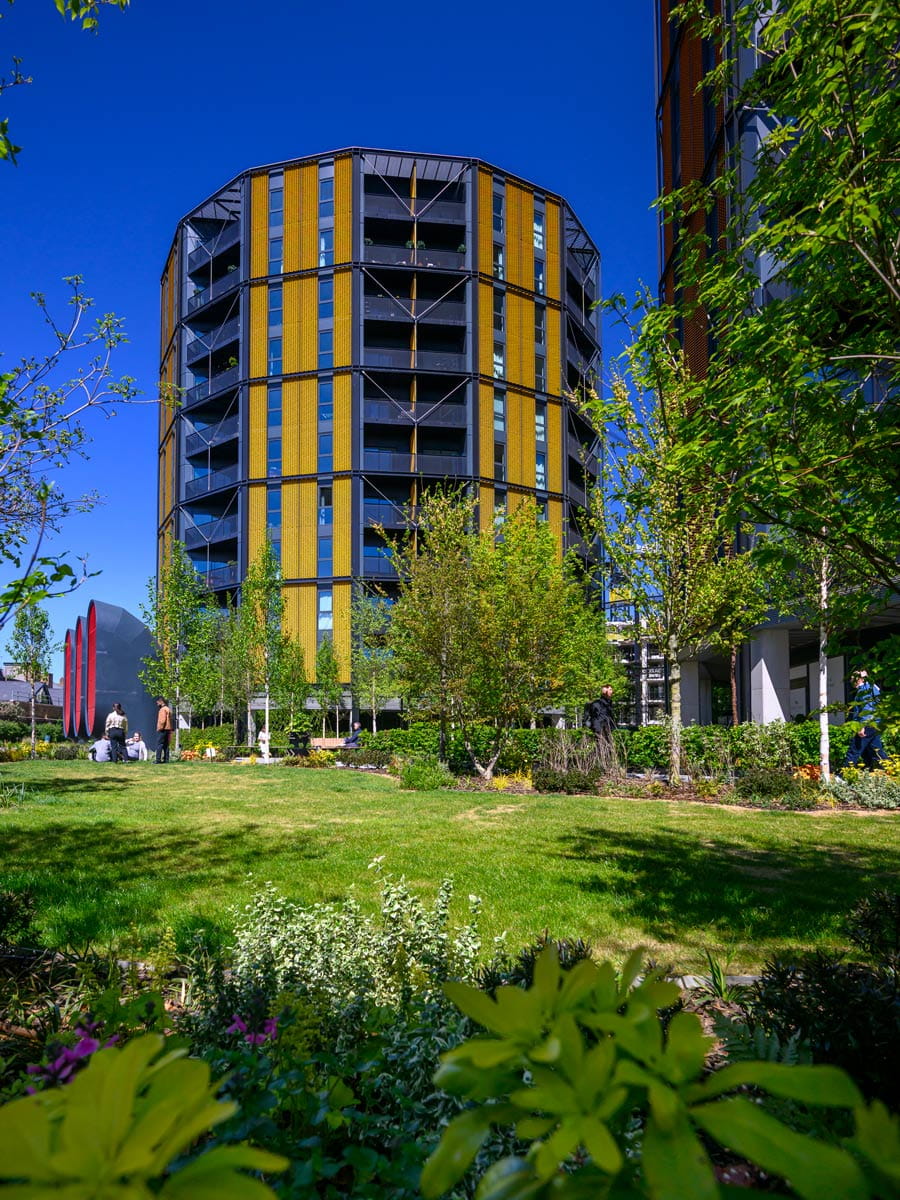 Modern yellow-and-grey apartment block beside a green lawn and trees under a clear blue sky.