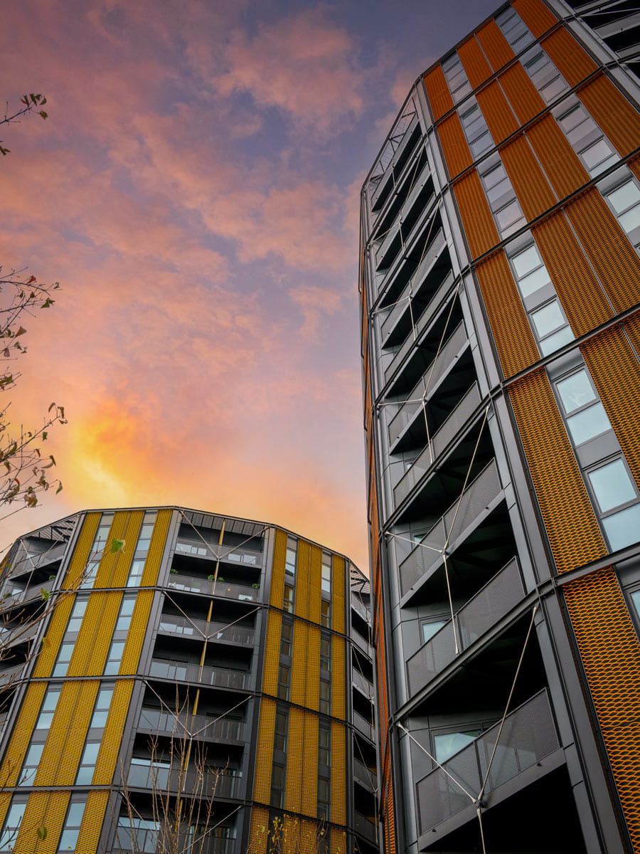 Low-angle view of modern flats with yellow cladding and balconies against a pink sunset sky.