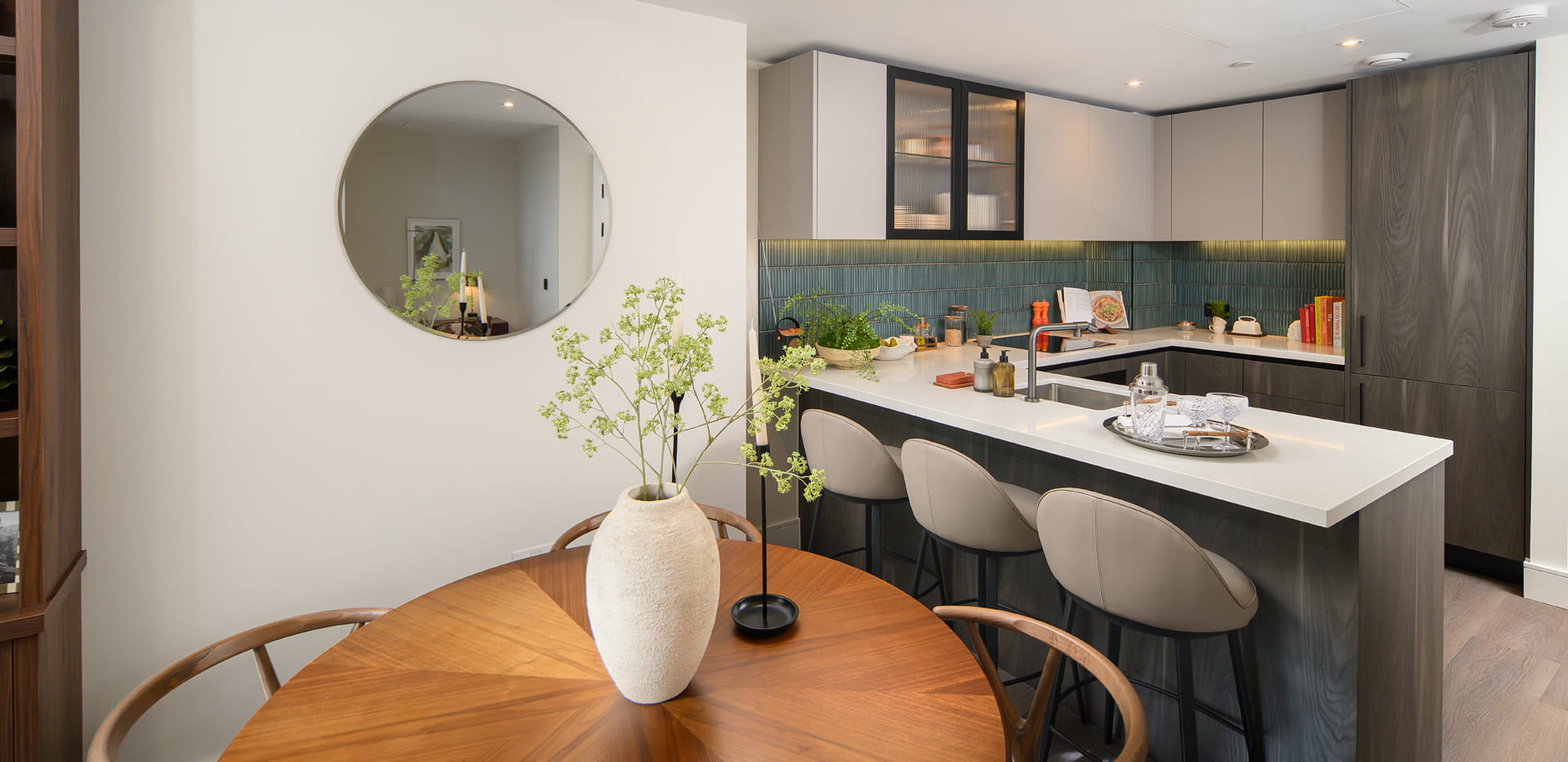 Wooden dining table in foreground, kitchen with island, dark green and grey tones.
