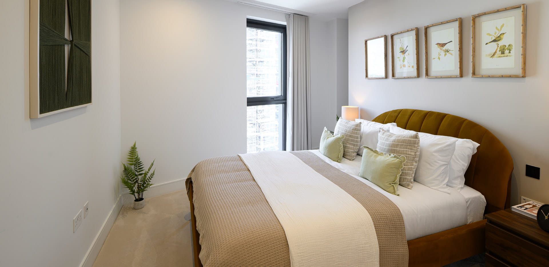 Modern bedroom with a double bed, framed bird prints, bedside table and a tall window with curtains.