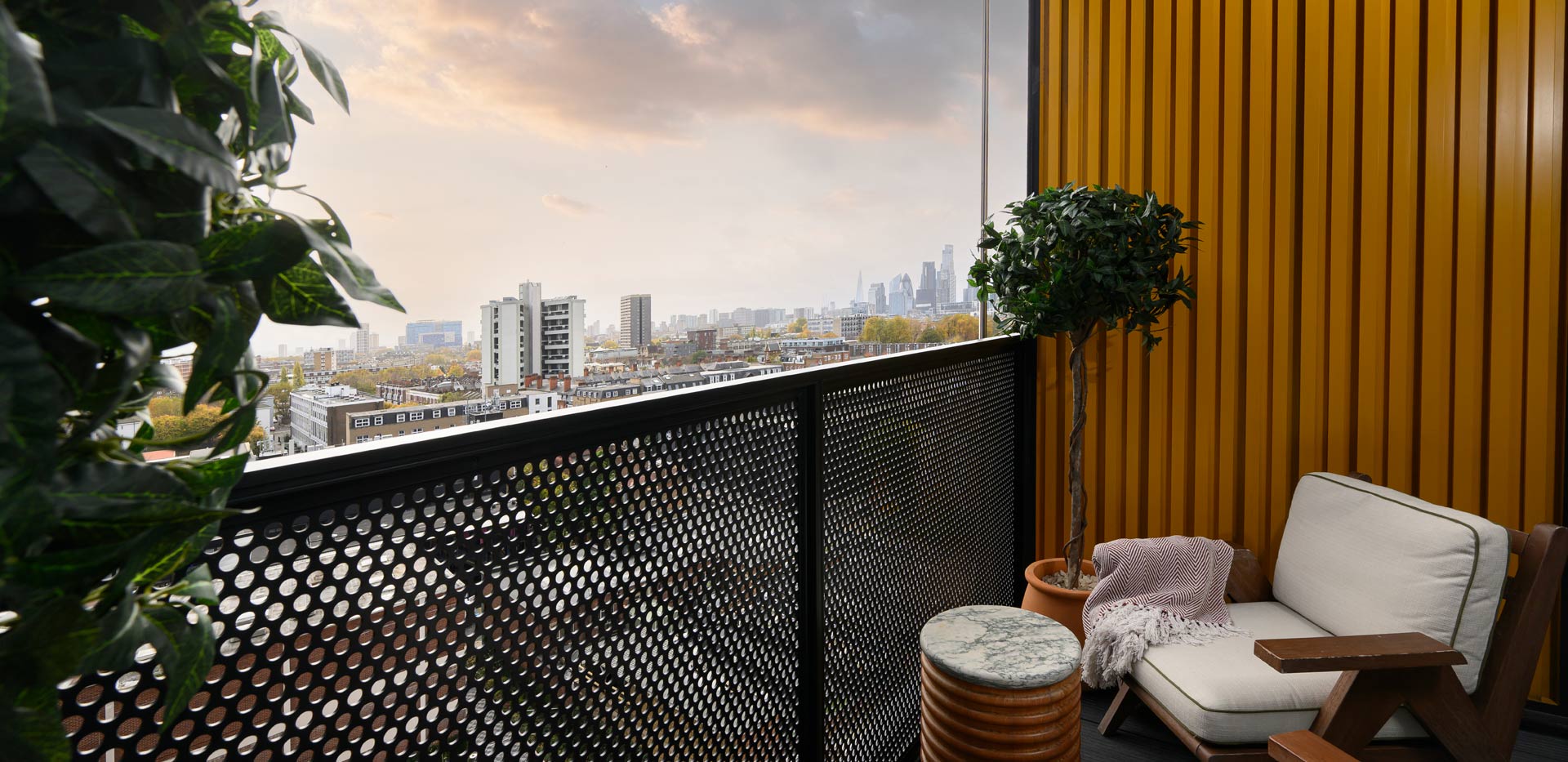 Modern balcony with cushioned chairs, potted plants and a city skyline view under a cloudy sky.