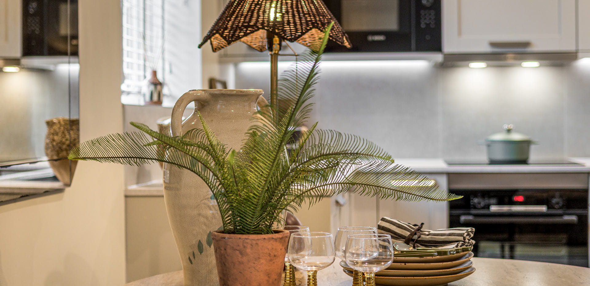 Closeup of dining table showing fern pot plant, glasses, cutlery, plates, ceramic vase and lamp