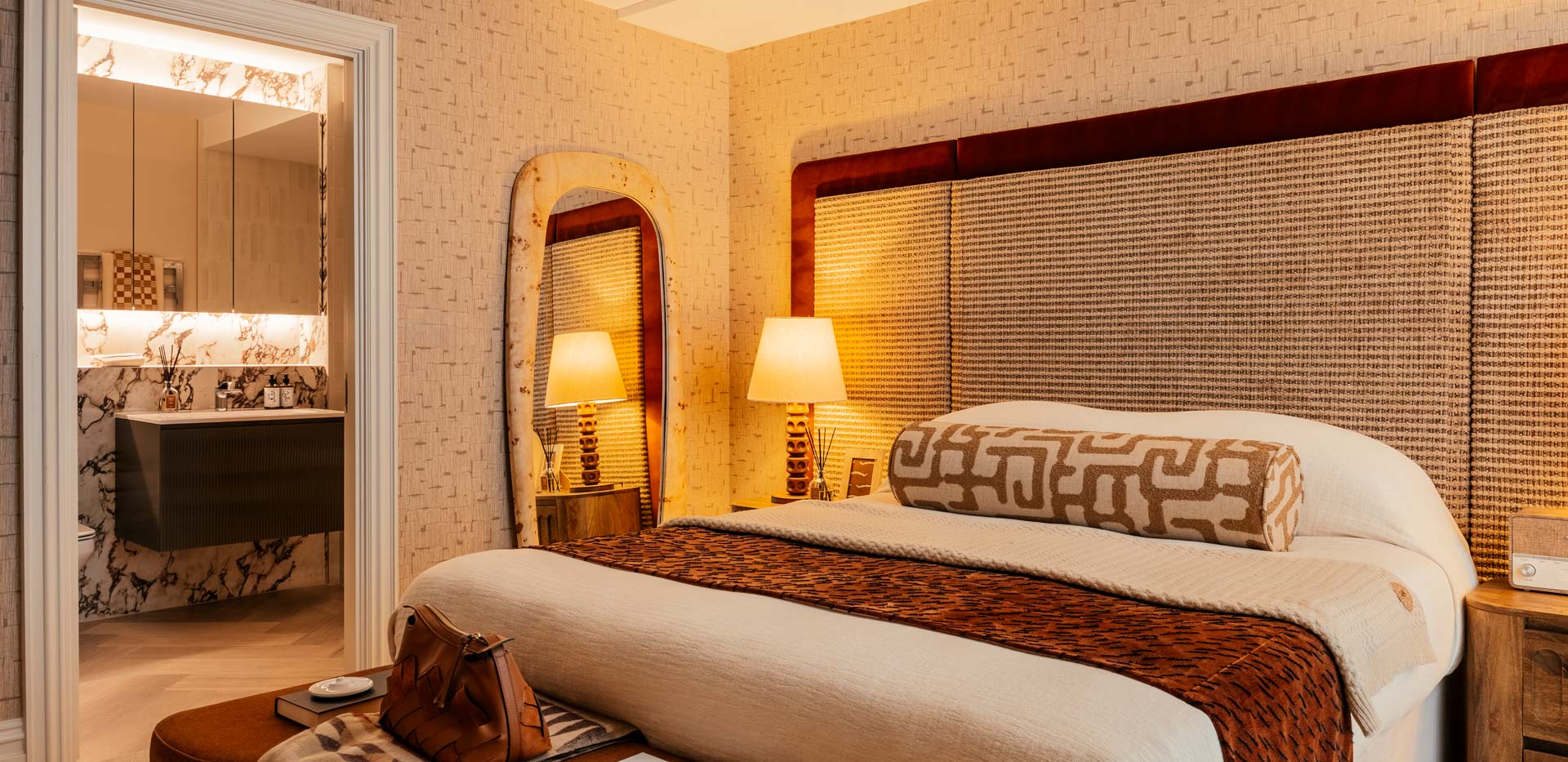 Double bed with tall and wide headboard, wall mirror, ottoman, door leading to ensuite bathroom.