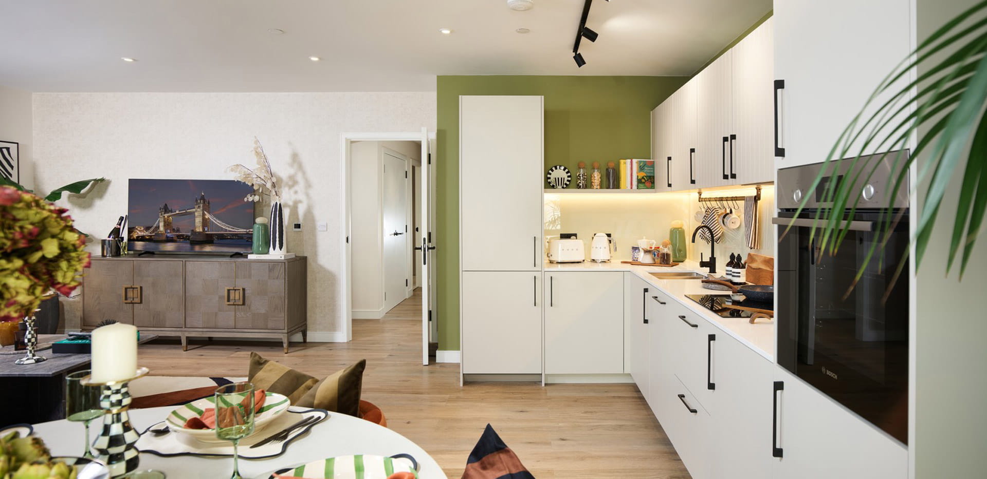 Decorated dining table in foreground, kitchenette with white panels and modern comfort appliances, in background.