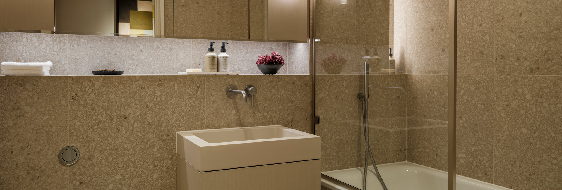 Modern designed bathroom with shower unit, beige-tan spotted tiles, wash basin with wall mirror and shelf space.