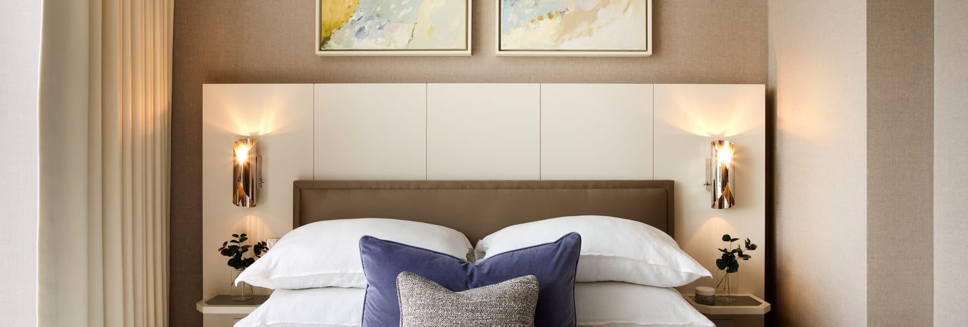 View of a made-up bed, padded cream-white headboard, bedside tables, wall hanging.