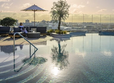 Sunlight reflecting of the shallow end of an outdoor swimming pool, deckchairs and cityscape view in background.
