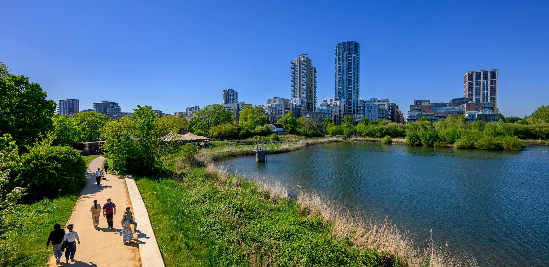 Pedestrians along a lakeside path, surrounded by greenery and tall residential buildings in background.