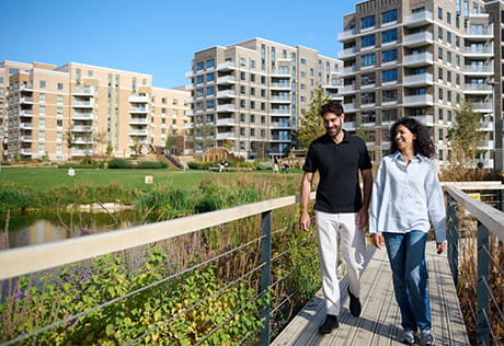 Two people on a boardwalk overlooking a water feature, grassed area, with new build apartment blocks in the background.