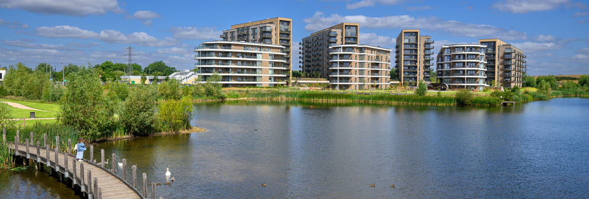 Calm lake with boardwalk pedestrian bridge in foreground, new build apartment blocks in the background